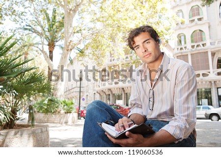 Young attractive businessman using a hands free device to make a call on his cell phone while sitting on a bench in a city square with classic architecture, smiling.