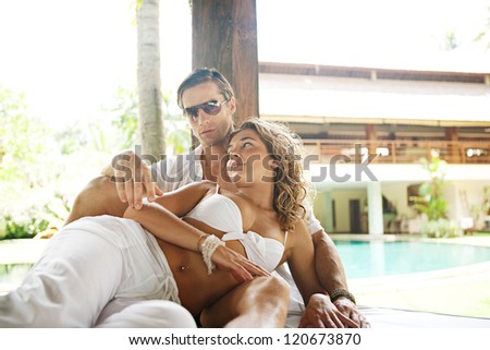 Young and attractive sexy couple lounging on a tropical garden bed in an exotic hotel spa garden, relaxing next to a swimming pool.