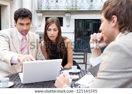 Three business people sharing a table at a coffee shop terrace, having a meeting and talking while using technology in the financial city district.
