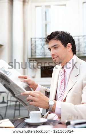 Attractive young businessman reading the newspaper while having breakfast at a coffee shop near classic office buildings in the city.