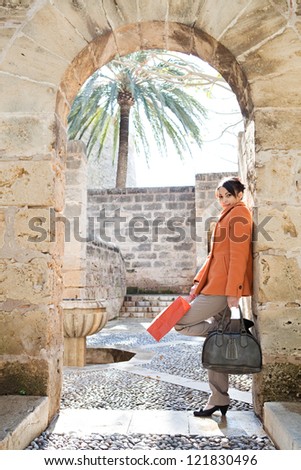 Attractive hispanic young woman leaning on an old stone wall while visiting a sight on vacations, holding shopping bags and smiling.
