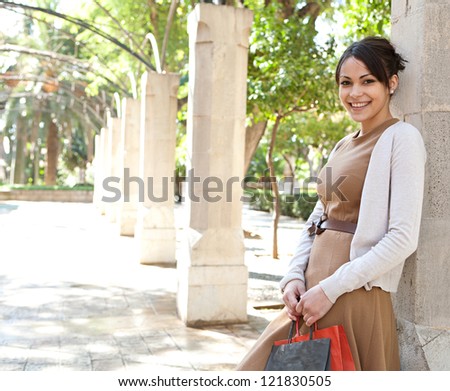 Portrait of an attractive young woman holding her shopping bags while leaning on a column in a romantic garden, outdoors.