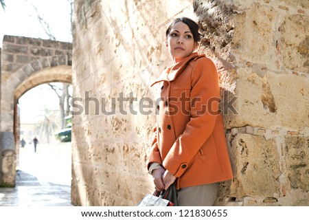 Attractive hispanic young woman leaning on an old stone wall while visiting a sight on vacations, holding shopping bags and being thoughtful.