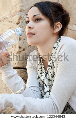 Young woman drinking water from a plastic blue bottle while sitting down near an old stone wall, outdoors.