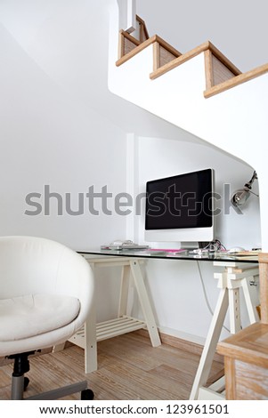 View of an empty modern home office space under wooden stairs, with a blank desktop computer screen.