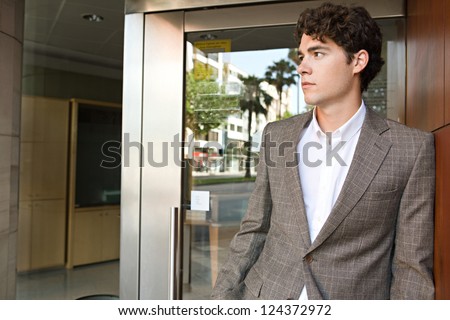 Young attractive businessman standing by an office building entrance, wearing a smart suit and looking away.