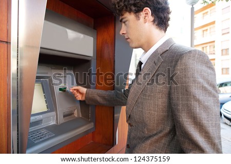 Side view of an elegant businessman withdrawing money from a wood decorated bank cash point, outdoors.