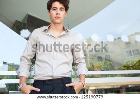 Graphic portrait of a confident businessman standing with his hands in his pockets by a modern building window with reflections of the city  behind him.