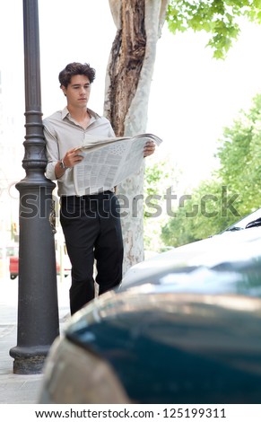 Young businessman leaning on an iron lamp post in the city, reading the newspaper with traffic and cars.