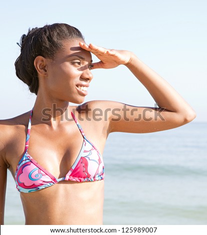 Attractive black woman covering her eyes to look at the horizon while standing in a colorful bikini on the beach on vacation.