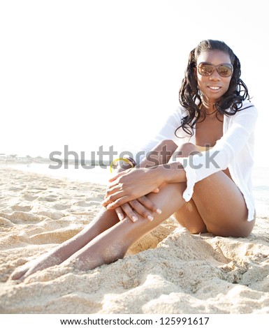 Young attractive black woman sitting on a white sand beach, relaxing while on vacation and smiling on a sunny day.