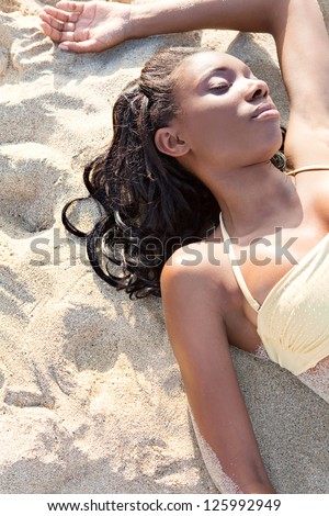 Sophisticated young african american woman laying down on a white sand beach, sunbathing wit her eyes closed, relaxing on holiday.