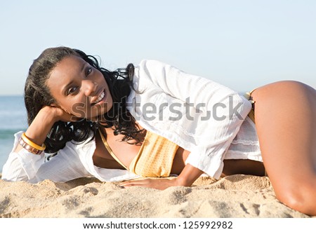 Attractive african american woman laying down on a golden sand beach by the sea shore, relaxing and sunbathing during a summer day.