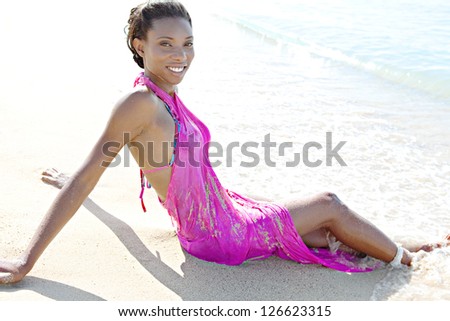 Side view of a beautiful black woman sunbathing in a beach shore, wearing a bright pink sarong with her feet in the water, on vacation.