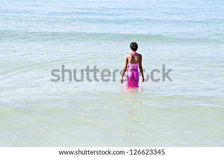 Black woman on a beach, walking into the sea wearing a bright pink sarong around her body while on vacations.