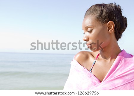 Close up profile portrait of an attractive black woman covering her shoulders with a pink sarong on the beach, being thoughtful against a blue sky.
