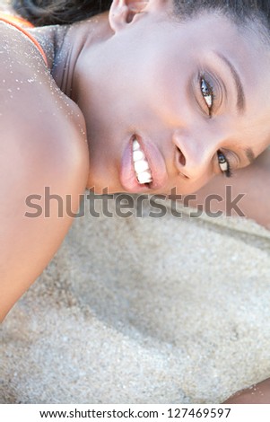 Over head close up beauty portrait of an attractive black woman with perfect skin, relaxing on a white sand beach, smiling.