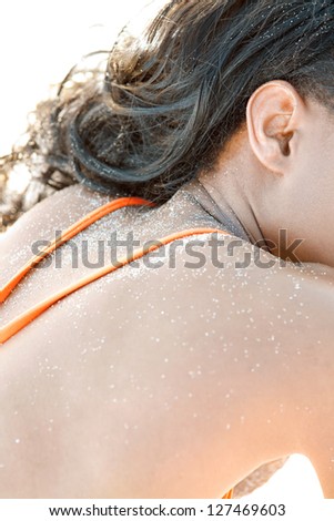 Rear part view of a woman back, shoulders and neck sunbathing on a white sand beach with grains on her skin, wearing an orange bikini.