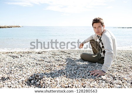 Confident smart man sitting down on the shore of a winter pebble beach relaxing and being thoughtful with a sunny blue sky in the background.