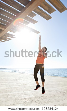 Figure of a sports man jumping up and reaching a wooden structure by the sea with the sun rays filtering through, against an intense blue sky.