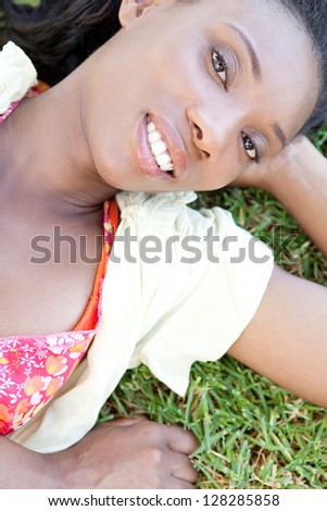Over head close up portrait of an attractive african american woman laying down on green grass, smiling outdoors.
