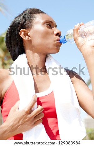 Portrait of an african american sport woman standing after doing sport with a towel around her neck, drinking mineral water from a bottle against a blue sky.