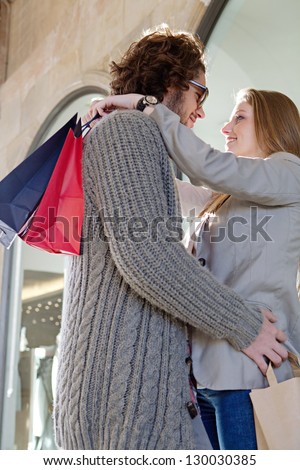 Young attractive tourist couple hugging outside a store window  with paper shopping bags, while on vacation, smiling.