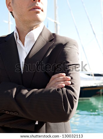 Middle section of a smart businessman wearing a suit and standing by a luxury yachts marine with his arms crossed against a blue sky.