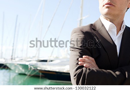 Close up middle section of a businessman standing by a luxurious yachts marine with his arms crossed against a deep blue sky and sea.