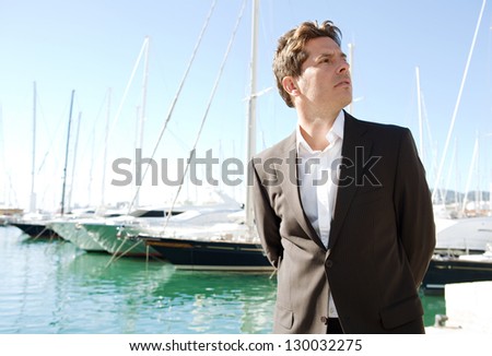 Close up portrait of an elegant and powerful businessman standing by a marine with luxury sailing boats moored in the background against a blue sky.