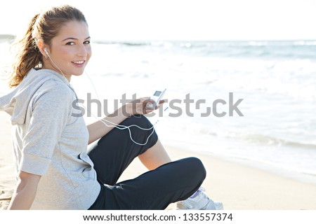 Portrait of a young woman sitting on a white sand beach shore, holding a music player and listening to music with her head phones, turning and smiling at the camera.