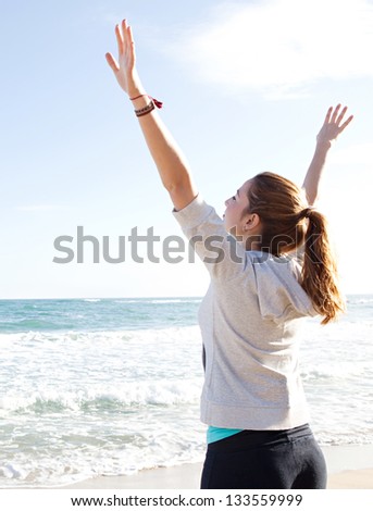 Side rear view of a young sporty woman stretching her arms up while standing on a beach by the shore and against a blue sky.
