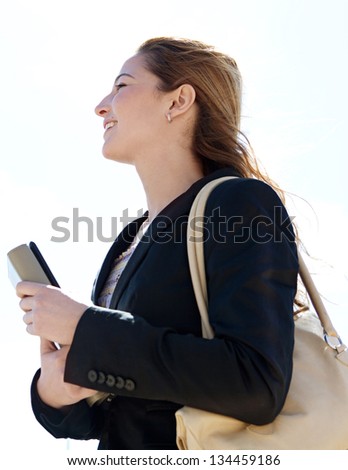 Profile portrait view of a young successful businesswoman carrying work folders and wearing a black suit, looking ahead against a blue sky, smiling.