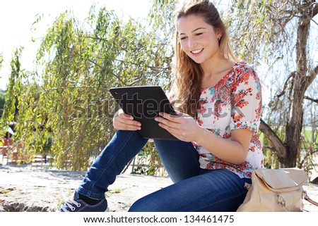 Young attractive woman using a digital tablet and touching the screen while sitting on a stone wall high up in a park with a blue sky during a sunny day.