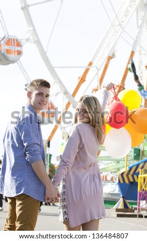 Attractive teenage couple in an amusement park with rides, holding balloons up in the air and turning around to look at the camera while holding hands.