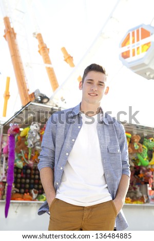Young teenager standing in an attractions park near the ferris wheel with games and toy prices behind him during a sunny day.