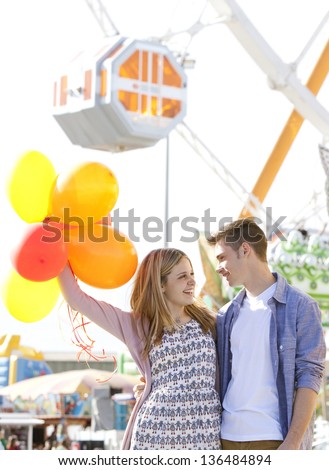 Young joyful couple having fun in a fun fair ground, smiling and holding colorful balloons up and holding each other.