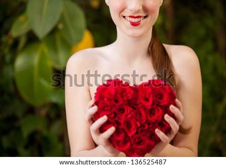 Attractive young woman naked in a green lush garden holding a red roses heart in front of her chest, wearing red lipstick, smiling.