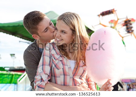 Close up of an attractive young couple kissing while visiting a funfair amusement park arcade with rides and holding a pink cotton candy floss sweet.