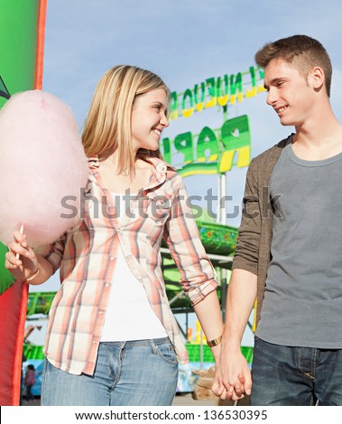 Young couple walking holding hands in an attractions park arcade with cotton candy floss and rides in the background.