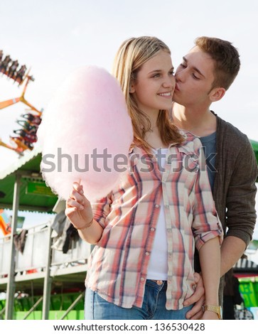 Young couple walking and kissing in an attractions park arcade with cotton candy floss and colorful rides in the background.