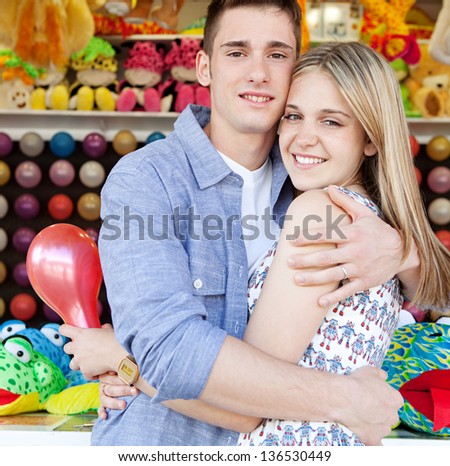 Portrait of a young teenage couple hugging while having fun in an amusement park arcade with colorful games and prices, holding a balloon and smiling.
