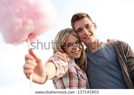 Close up portrait of a young attractive couple in a fun fair, holding a cotton candy sweet towards the camera, smiling against a blue sky.