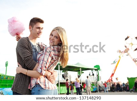 Attractive young couple hugging and having fun in an amusement park arcade during a sunny day, holding a cotton candy sweet and turning to the camera smiling.