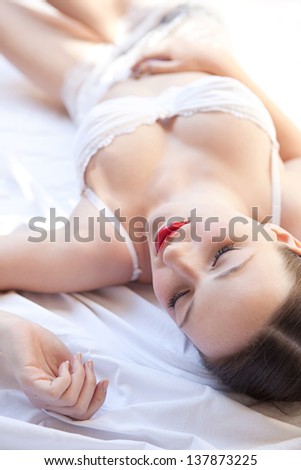 Over head beauty portrait of a young woman laying down in bed wearing white lingerie and red lips, sleeping.