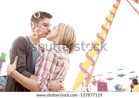 Side portrait view of a young couple kissing while visiting an attractions park arcade with bubbles floating around them against the sky.