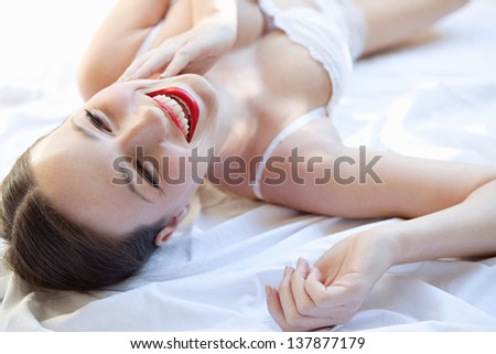 Over head beauty portrait of a young woman laying down in bed wearing white lingerie and red lips, smiling.