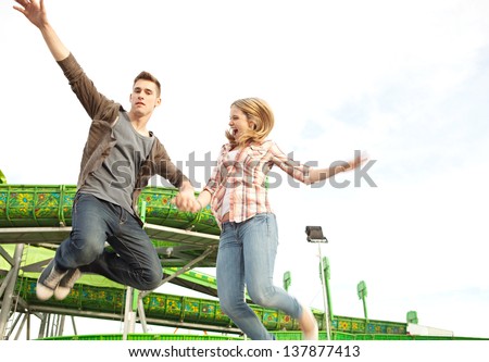 Teenage couple holding hands and jumping up in the air next to an attractions ride in an amusement park arcade during a sunny day.