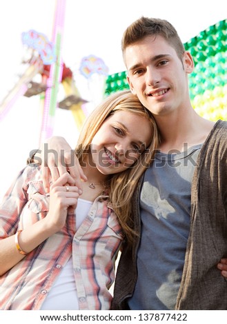 Joyful young couple holding each other while visiting an attractions park arcade with a ticket office and rides in the background.