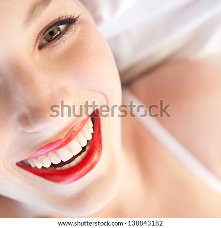 Over head beauty portrait of a young woman\'s half face laying down in bed wearing white lingerie and red lips, smiling at the camera.